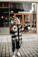 un elegante mujer soportes en frente de un edificio, agarrando un vibrante rojo café taza. su negro Saco y tartán bufanda añadir instinto a el urbano escena, capturar el esencia de un enérgico ciudad Mañana foto
