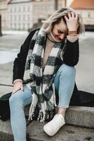 A woman in a plaid scarf and jeans sits on steps, smiling warmly at the camera. The relaxed atmosphere captures a moment of genuine happiness and comfort. Casual Charm. Smiling Woman on Steps photo