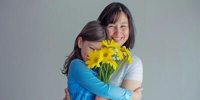 Adorable young girl in blue embraces her mother, who holds yellow flowers, with a studio-lit gray wall behind. photo