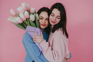 madre abraza hija en contra un rosado fondo. mujer en azul suéter participación tulipanes, vestido en pastel rosado foto