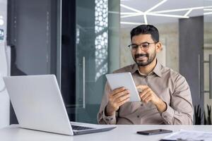 A cheerful Indian programmer is smiling and using a digital tablet, sitting at his desk in a modern office setting. photo