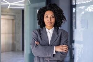Portrait of serious successful African American business woman, boss in business suit looking focused at camera with crossed arms, female financial worker inside office at workplace. photo