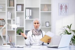 Professional woman in hijab and lab coat in a clinic office, engaging in patient consultation with laptop and medical equipment. photo