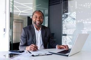 Portrait of successful mature experienced financier, business man behind paper work smiling and looking at camera, african american man at workplace reviewing contracts, accounts and reports. photo