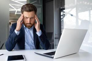 Stressed businessman with headache at office desk, feeling overwhelmed and anxious, work stress concept photo