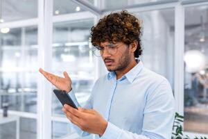 Close up of businessman in middle of office holding phone, man upset reading bad news online, hispanic man disappointed and depressed by notification. photo