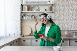 Beautiful young woman in glasses and green shirt relaxing at home in kitchen, listening music photo