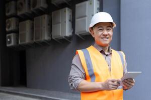 retrato de un sonriente joven masculino construcción trabajador en un difícil sombrero y chaleco participación un tableta en su manos y mirando a el cámara. foto