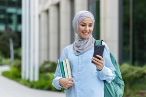 joven musulmán mujer en hijab caminando fuera de Universidad instalaciones, hembra estudiante sonriente contentamente utilizando aplicación en teléfono, mochila en espalda y libros en manos. foto