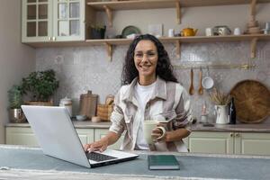 retrato de joven hermosa Hispano mujer a hogar, hembra persona de libre dedicación trabajando remotamente utilizando computadora portátil, mirando a cámara y sonriente en lentes y Rizado pelo en cocina. foto