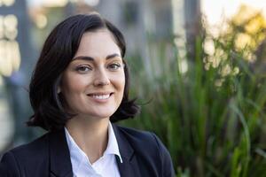 Portrait of a confident businesswoman smiling warmly, dressed in professional attire, standing outdoors with a soft focus background of greenery. photo