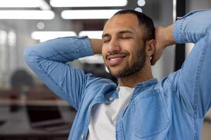 un de cerca foto de un joven latín americano hombre sentado contentamente y sonriente en un moderno oficina con su ojos cerrado y su brazos, relajado detrás su cabeza.