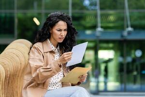 A worried young woman is sitting on a bench outside, intently reading a document with a serious expression. photo