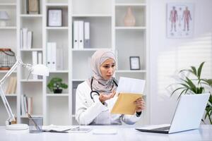 un enfocado musulmán hembra médico en un hijab y blanco Saco examina documentos en su médico oficina, simbolizando cuidado de la salud diversidad. foto