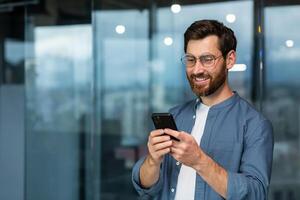 exitoso empresario en lentes y barba en pie cerca ventana en el noche, hombre utilizando teléfono, contento jefe mecanografía mensaje y hojeada en línea paginas en teléfono inteligente aplicación dentro oficina. foto