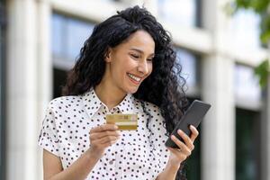 joven hermosa mujer caminando en el ciudad, satisfecho latín americano mujer con Rizado pelo sostiene teléfono y banco crédito tarjeta en manos, hace en línea compras felizmente libros servicios y elige en línea. foto