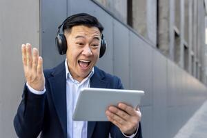 Happy young Asian male businessman standing in headphones outside office center, looking at tablet screen and happy with news and success. photo
