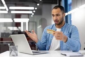 A man is holding a credit card in front of a laptop computer. He looks surprised and is possibly questioning the validity of the card photo