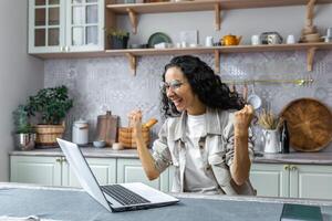 emocionado y contento Hispano mujer celebrando victorioso el lotería por levantamiento su manos. sentado a hogar en el cocina con un computadora portátil, recepción un nuevo trabajo oportunidad, recibido bueno noticias. foto