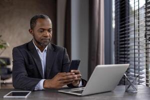 Focused African American businessman multitasking with smartphone and laptop at a modern office workspace, exuding professionalism and efficiency. photo
