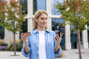 Happy successful woman with phone and bank credit debit card in hands, choosing gifts in online store, joyfully booking services and transferring money while standing outside office building. photo