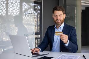 un sonriente joven hombre trabajos en el oficina a un computadora portátil, tipos en un teclado y usos un crédito tarjeta en su manos, conduce en línea actas, cheques un cuenta. foto