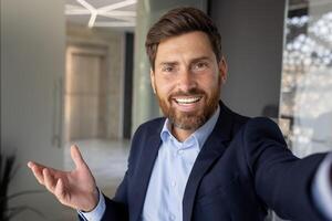 Remote call conversation in the office. A young businessman is standing in a suit and holding a phone, looking at the camera and smiling. Close-up photo. photo