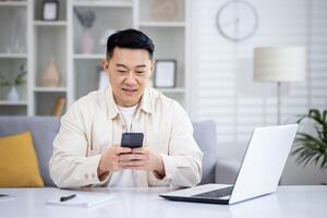 Mature Asian man sitting at table at home, man using phone, typing message on smartphone, app user and browsing internet in home cinema room, smiling contentedly. photo