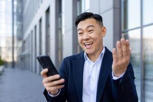 Close-up photo of a young Asian male businessman standing outside an office center, looking at the phone screen and emotionally happy.