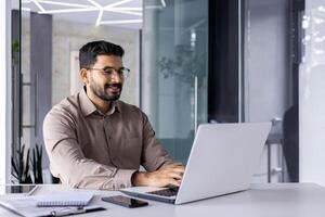 empresario contento exitoso y sonriente trabajando con ordenador portátil dentro oficina a lugar de trabajo, hombre satisfecho con financiero resultado prepara informe, contador financiero mecanografía en computadora teclado. foto