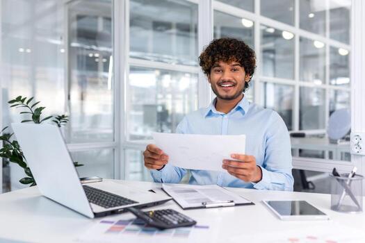 retrato de joven exitoso Hispano empresario dentro oficina, hombre sonriente y mirando a cámara, papel trabajador contento con logro resultados sentado a lugar de trabajo con ordenador portátil. foto