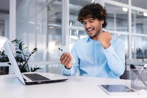 Young successful businessman inside the office at work uses a laptop hispanic man holds a smartphone in his hands, the man holds his hand up a gesture of success and triumph, celebrating victory. photo