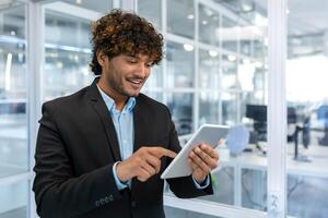 joven exitoso empresario con tableta computadora , sonriente Hispano hombre trabajando dentro un moderno oficina edificio a trabajo foto