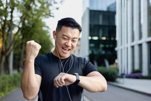 de cerca foto. un joven asiático hombre lo hace Deportes, carreras en el ciudad calle en auriculares, mira a el resultado en un inteligente mirar, se regocija, demostración un victoria gesto con su mano. foto