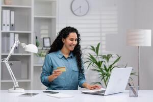 Focused Hispanic businesswoman using laptop and credit card in a modern home office setting, exemplifying remote work and e-commerce. photo