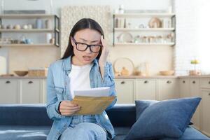 Shocked and alarmed young beautiful Asian woman received divorce papers. Sitting at home on the couch and holding an envelope with papers in his hands. Holding his head from unexpected news. photo