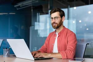 Serious and focused mature businessman in red shirt wearing glasses looking at camera, young man at work typing on laptop keyboard, programmer developer portrait at work. photo