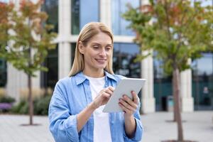 Smiling blonde woman using digital tablet while standing outdoors at pedestrian space with trees. Confident landscape designer developing green zone area in district with futuristic skyscrapers. photo