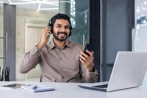 Carefree hispanic man in headphones holding cellular phone in hand while sitting by equipped desktop in office. Relaxed manager fixing wireless accessory for listening to music during workday. photo