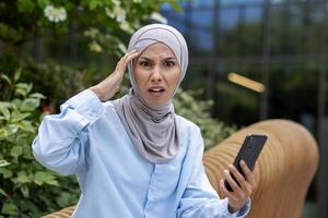 joven árabe mujer en hijab muestra un sorprendido expresión mientras comprobación teléfono inteligente fuera de moderno oficina edificio en urbano ajuste. foto