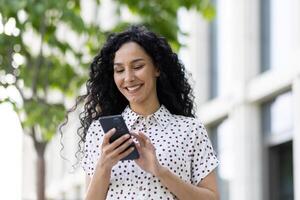 alegre mujer con Rizado pelo utilizando teléfono inteligente al aire libre. ella exhibiciones felicidad y conectividad en un casual urbano ajuste. foto