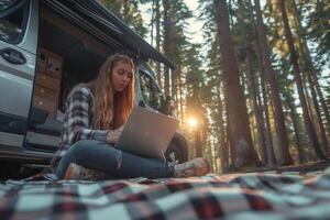 A young female digital nomad is using her laptop in the forest sitting on a mat near her travel van photo