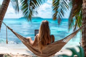 back view of a young woman sitting on a hammock facing the sea operating a laptop photo