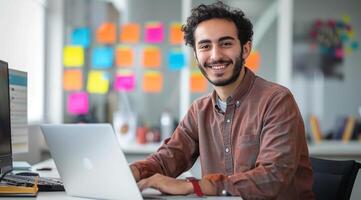 Portrait of a smiling young Arabian man sitting at a desk with a laptop in a modern office photo