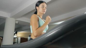 Young athletic woman is running on the treadmill at the fitness club. Concentrated young girl jogging in the underground gym. Low-angle view video