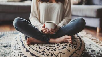 un persona sentado con las piernas cruzadas en un amortiguar practicando atención plena con un taza de café en mano tomando en cada sorbo con intención y gratitud foto