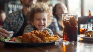 un niñito felizmente masticando en un pollo licitaciones plato mientras su padres disfrutar un frío soda a el bar foto