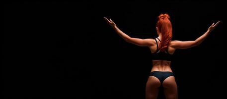 athletic redhead girl in sports underwear on a black background, rear view banner photo