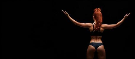 athletic redhead girl in sports underwear on a black background, rear view banner photo