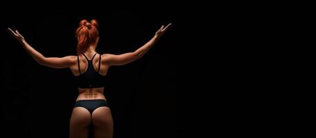 athletic redhead girl in sports underwear on a black background, rear view banner photo